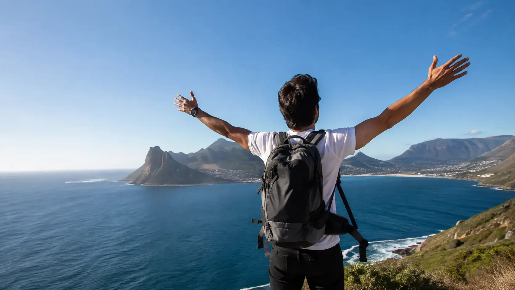 Man with a backpack standing arms open above a coastal mountain bay